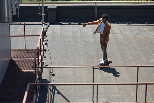 stylish african american businessman riding longboard on sunny rooftop