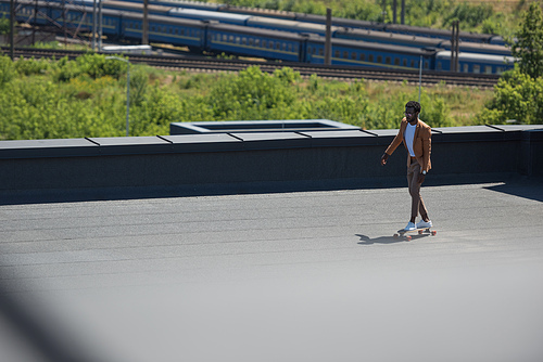 selective focus of african american businessman riding longboard on sunny rooftop with railways and trains on background
