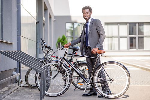 handsome african american businessman smiling at camera while taking bike from parking