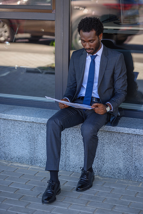 focused african american businessman reading newspaper while sitting near office building