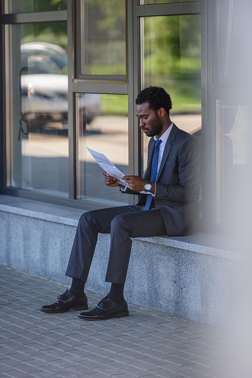 selective focus of attentive african american businessman reading newspaper while sitting near office building