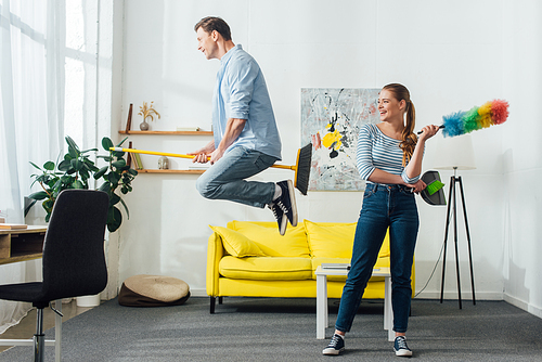 Smiling woman with dust brush and scoop looking at boyfriend levitating on broom in living room