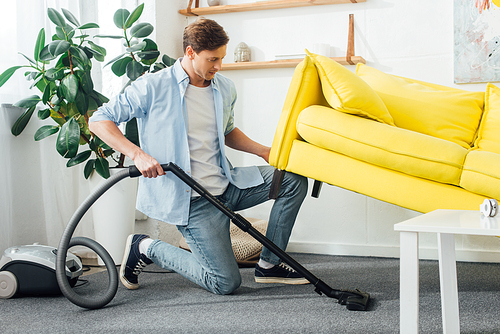 Side view of man lifting up couch while cleaning carpet with vacuum cleaner