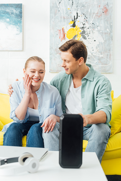 Selective focus of man embracing smiling girlfriend on couch near wireless speaker and headphones on coffee table
