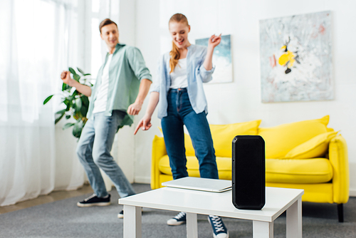Selective focus of wireless speaker and laptop on coffee table and cheerful couple dancing at home