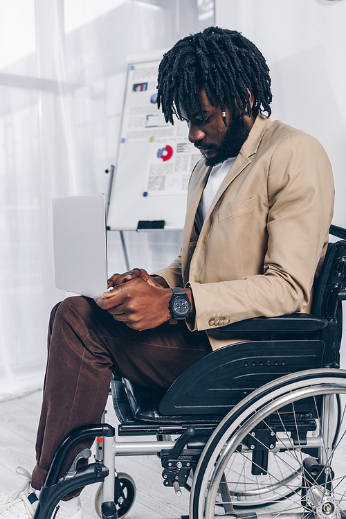 Selective focus of african american disabled employee on wheelchair working on laptop in office