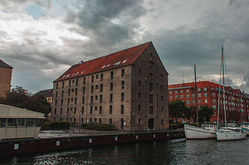 Buildings near harbor and boats on water with cloudy sky at background in Copenhagen, Denmark
