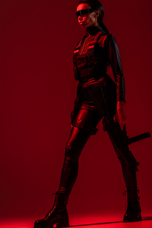 futuristic african american woman in glasses with walking assault rifle on red background