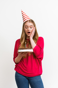 surprised girl in party cap looking at birthday cake isolated on white