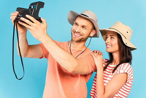 happy man in hat holding vintage camera and taking selfie with cheerful girl isolated on blue