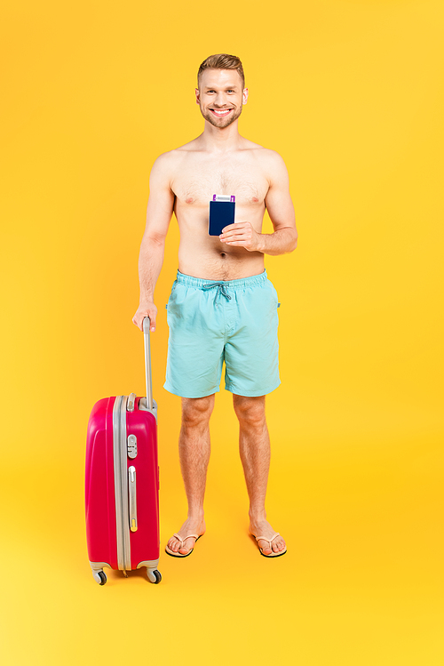 happy and muscular man holding passport and standing with baggage on yellow