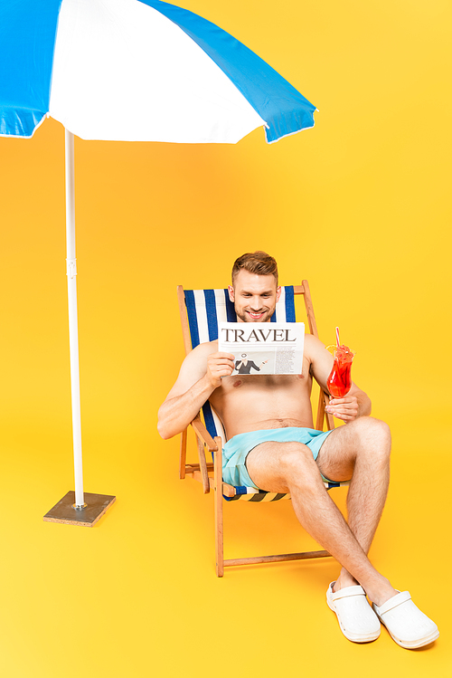 handsome man sitting on deck chair with cocktail while reading travel newspaper near beach umbrella on yellow