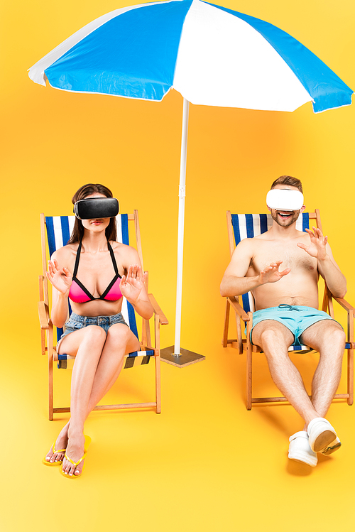 happy couple in virtual reality headsets sitting on deck chairs and gesturing near beach umbrella on yellow