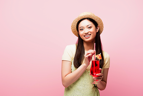 smiling asian woman in straw hat holding glass with cocktail isolated on pink