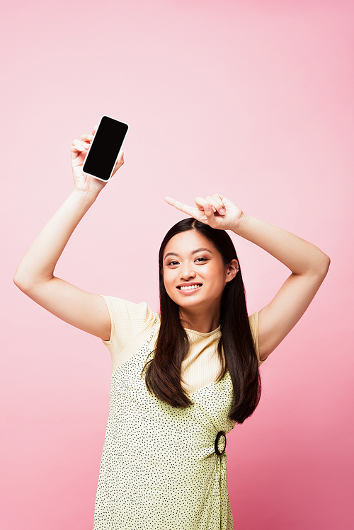 cheerful asian girl pointing with finger at smartphone with blank screen isolated on pink