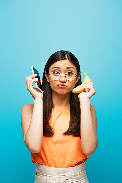 confused asian girl in glasses holding smartphone and banana isolated on blue