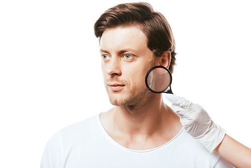 Young man looking away near dermatologist with magnifying glass isolated on white