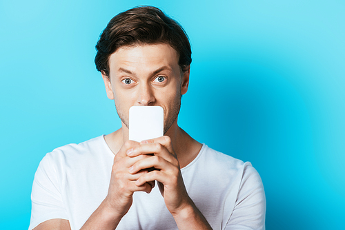 Young man covering mouth with smartphone on blue background