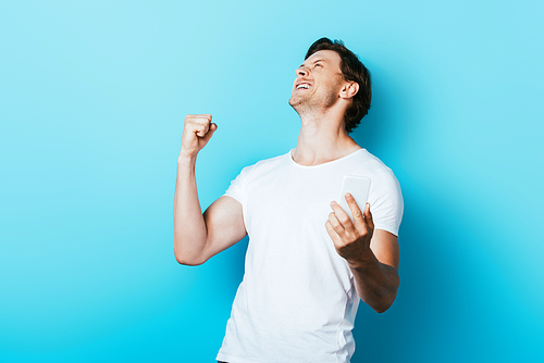 Young man showing yes gesture while holding smartphone on blue background