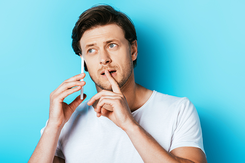 Thoughtful man with finger near lips talking on smartphone on blue background