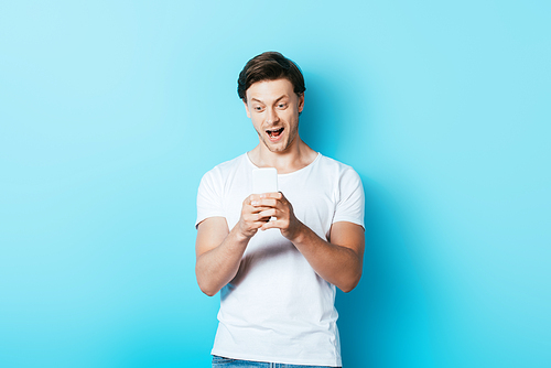 Excited man in white t-shirt using smartphone on blue background