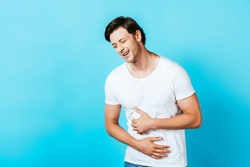 Young man in white t-shirt laughing and touching belly on blue background