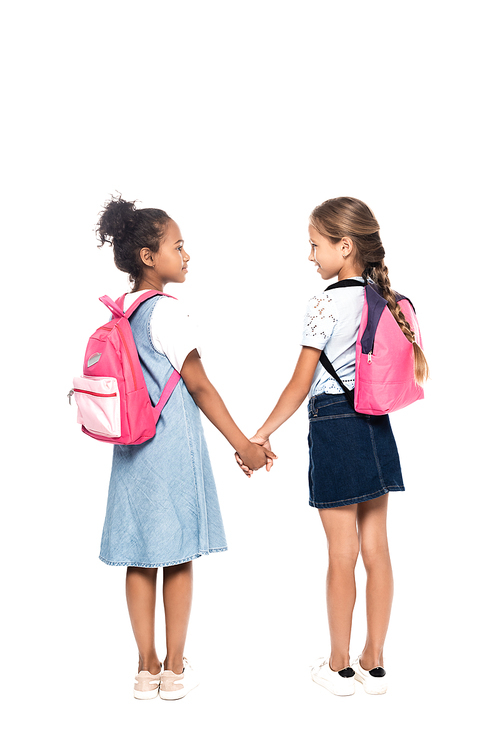 multicultural schoolkids with backpacks holding hands and looking at each other isolated on white