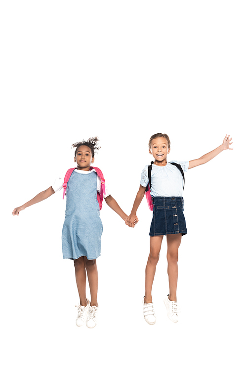 multicultural schoolgirls with backpacks holding hands and jumping isolated on white