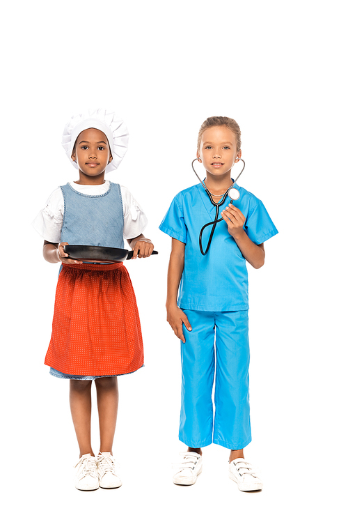 multicultural kids in costumes of different professions holding frying pan and stethoscope while standing isolated on white