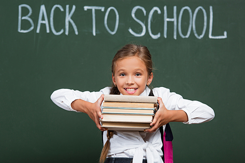 excited schoolgirl holding stack of books near chalkboard with back to school inscription