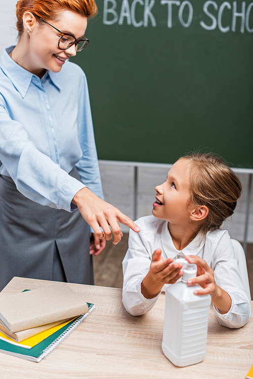 selective focus of smiling teacher pointing with finger near schoolgirl applying hand antiseptic