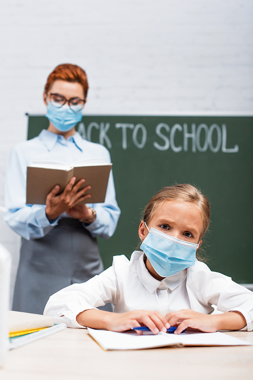 selective focus of bored schoolgirl in protective mask , and teacher standing near chalkboard with back to school lettering