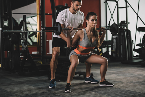 attentive, handsome trainer supporting young, attractive sportswoman lifting barbell