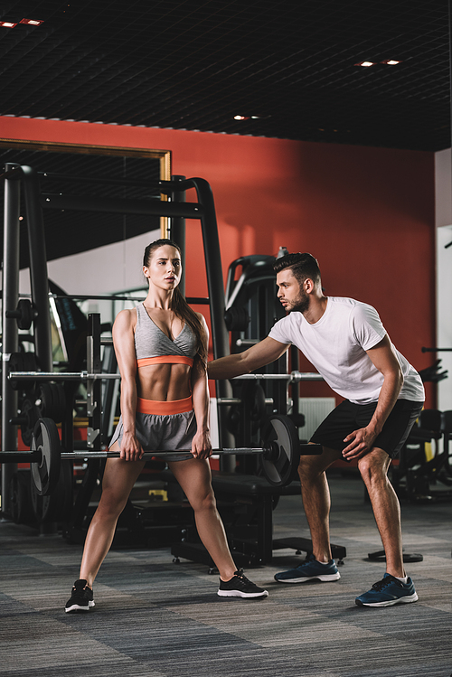 handsome trainer supporting attractive sportswoman lifting barbell