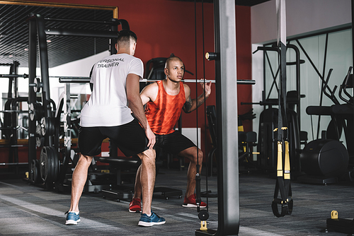 back view of personal trainer supervising young african american sportsman lifting barbell