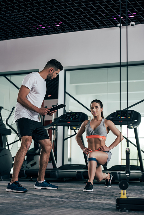 personal trainer holding clipboard while looking at attractive sportswoman exercising with resistance band