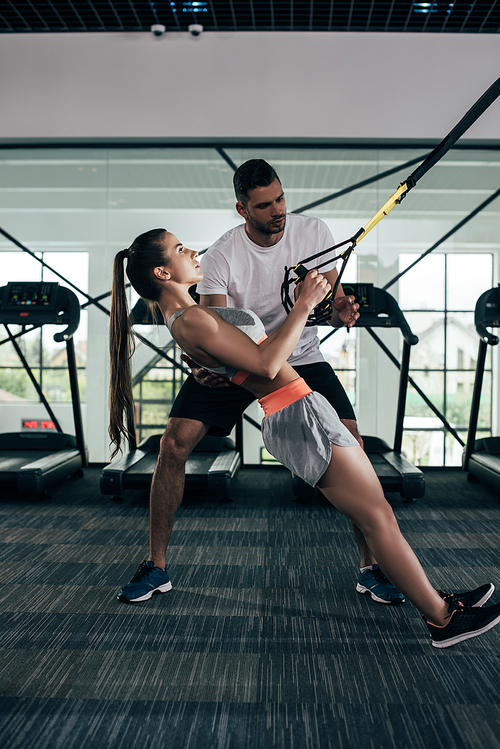 attentive trainer supporting young sportswoman working out on suspension trainer
