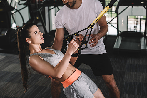 cropped view of trainer supporting young sportswoman pulling up on suspension trainer