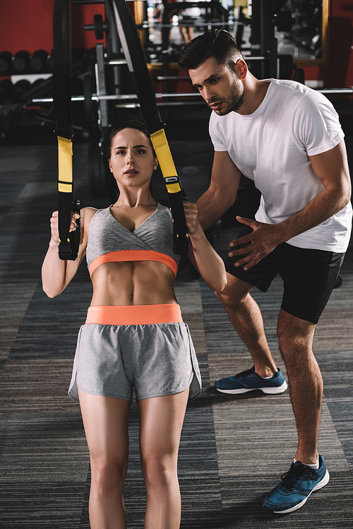 attentive trainer supporting young sportswoman pulling up on suspension trainer