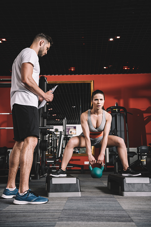 attentive trainer holding clipboard while standing near supervising young sportswoman lifting weight in gym