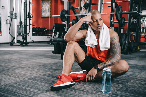 exhausted african american sportsman sitting on floor near sports bottle