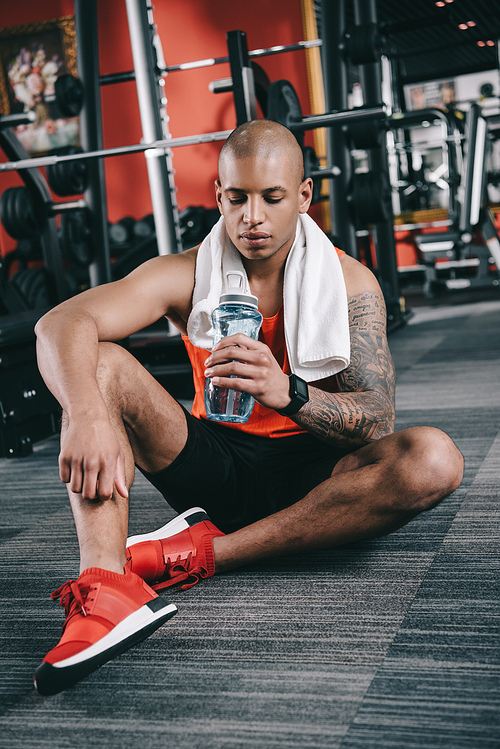 tired african american sportsman holding sports bottle while sitting on floor