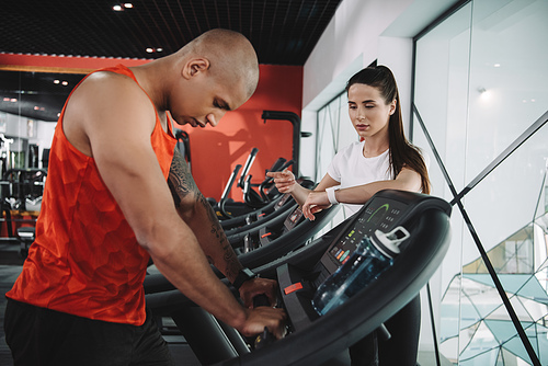 attentive trainer standing near african american sportsman running on treadmill