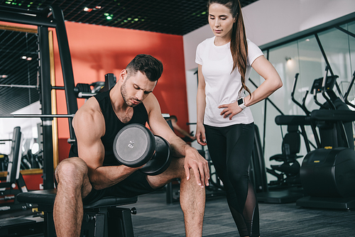 attentive trainer standing near handsome sportsman working out with dumbbell