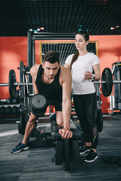 attentive trainer assisting handsome sportsman working out with dumbbell