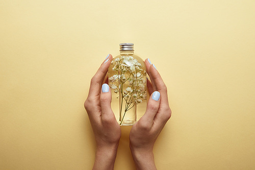 partial view of woman holding bottle with natural beauty product and herbs in hands on yellow background