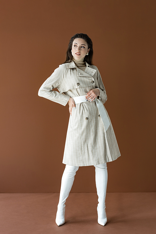 trendy woman in stylish trench coat isolated on brown with hand on hip