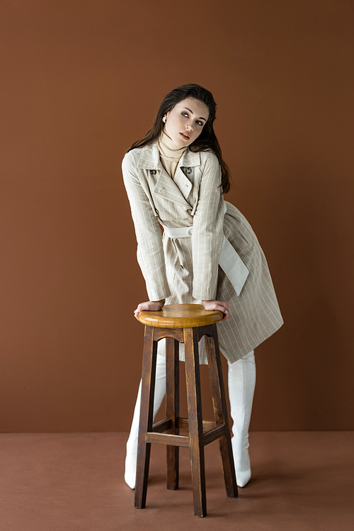 attractive adult woman standing near chair isolated on brown, looking away
