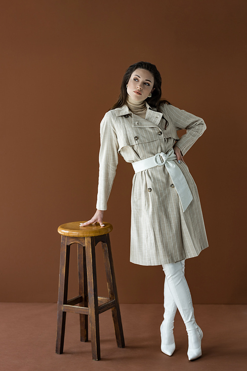 fashionable woman in stylish trench coat looking away, standing near chair isolated on brown