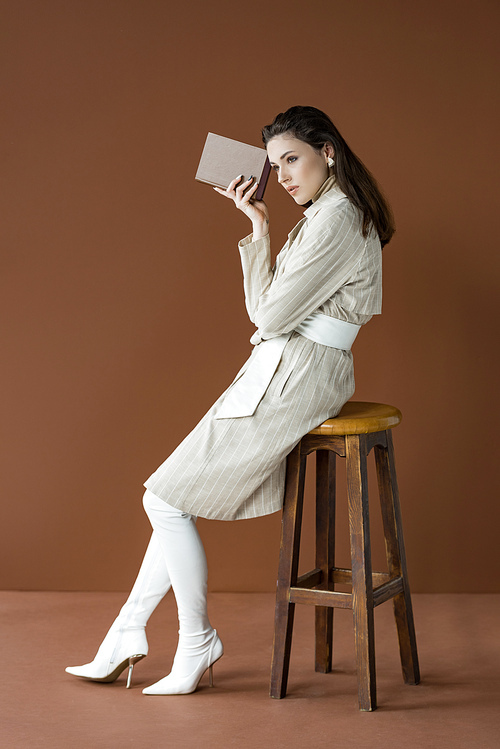 side view of beautiful stylish woman in trench coat holding book, sitting on chair isolated on brown